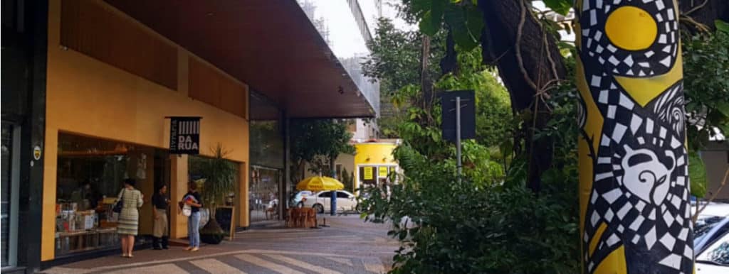 Uma vibrante cena de rua em Belo Horizonte com um prédio amarelo, arte na calçada, árvores e pessoas perto de uma loja com uma placa "DA RUA". Um poste colorido com padrão abstrato se destaca em primeiro plano, à direita.
