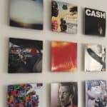 Nove capas de álbuns de vinil dispostas em uma grade 3x3 em uma parede branca criam uma decoração musical impressionante, apresentando vários designs coloridos e em preto e branco, incluindo um com a palavra “CASH” em letras maiúsculas.