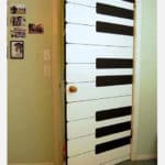 Uma porta pintada para imitar teclas de piano, com retângulos pretos e brancos alternados, acrescenta um toque de decoração musical. Há fotos e interruptores de luz na parede verde-claro adjacente.