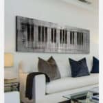 Uma sala de estar moderna com um sofá branco, almofadas claras e escuras, uma mesa lateral de vidro com um abajur e uma grande obra de arte de parede acima do sofá com teclas de piano abstratas em preto e branco para um toque de decoração musical.