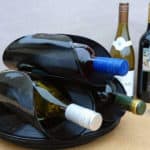 Duas garrafas de vinho são armazenadas em um suporte para vinhos feito de discos de vinil pretos reciclados, adicionando um toque de decoração musical. Ao fundo, três garrafas de vinho e duas taças vazias repousam sobre uma superfície bege com fundo neutro.