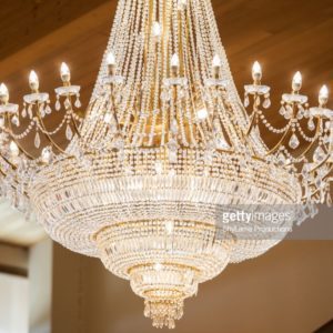 Um grande lustre de cristal ornamentado com v&aacute;rios n&iacute;veis e l&acirc;mpadas acesas semelhantes a velas pende do teto de madeira, brilhando &agrave; luz.