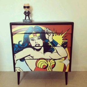 Uma c&ocirc;moda pintada com uma imagem colorida em estilo de hist&oacute;ria em quadrinhos da Mulher-Maravilha, com uma estatueta colecion&aacute;vel em cima da c&ocirc;moda, contra uma parede branca lisa.