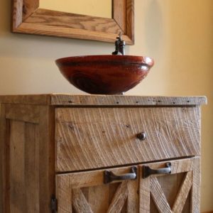 Uma penteadeira r&uacute;stica de madeira com uma porta de arm&aacute;rio estilo celeiro, encimada por uma pia redonda marrom-avermelhada e uma torneira simples. Acima dela, pende um espelho com moldura de madeira combinando. Um casti&ccedil;al est&aacute; fixado na parede pr&oacute;xima.