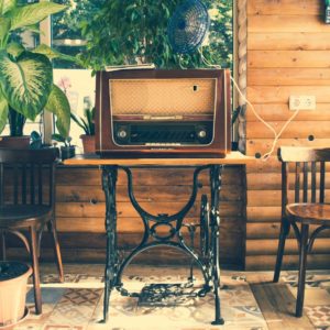 Um r&aacute;dio antigo repousa sobre uma pequena mesa de madeira com pernas de metal, entre duas cadeiras de madeira. H&aacute; vasos de plantas e paredes com pain&eacute;is de madeira em uma sala ensolarada com piso de ladrilhos estampados.