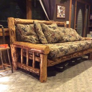 Um sof&aacute; r&uacute;stico de madeira com estrutura grossa em estilo tronco e almofadas estampadas est&aacute; situado em uma sala aconchegante com carpete bege, mesa lateral de madeira e grandes janelas cobertas por cortinas escuras. Caixas de armazenamento ficam embaixo do sof&aacute;.