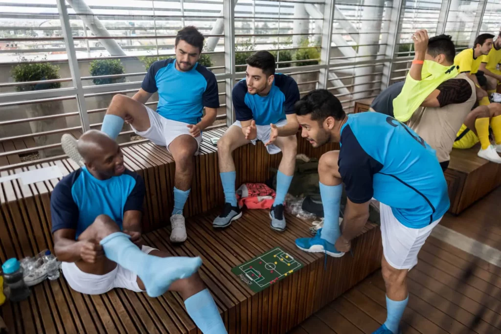 Quatro jogadores de futebol masculinos, de uniforme azul, sentam-se em bancos de madeira em um vestiário, preparando-se para uma partida e discutindo estratégias com um pequeno quadro tático, muito parecido com aqueles vistos antes dos jogos nos campos de futebol em São Paulo. Dois jogadores de amarelo sentam-se ao fundo.