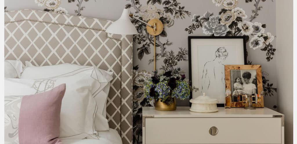Um quarto estiloso com cabeceira estampada, roupa de cama branca com almofada rosa e papel de parede floral é uma tendência em decoração. A mesa de cabeceira abriga flores, quadros e fotos, um abajur e um prato coberto.