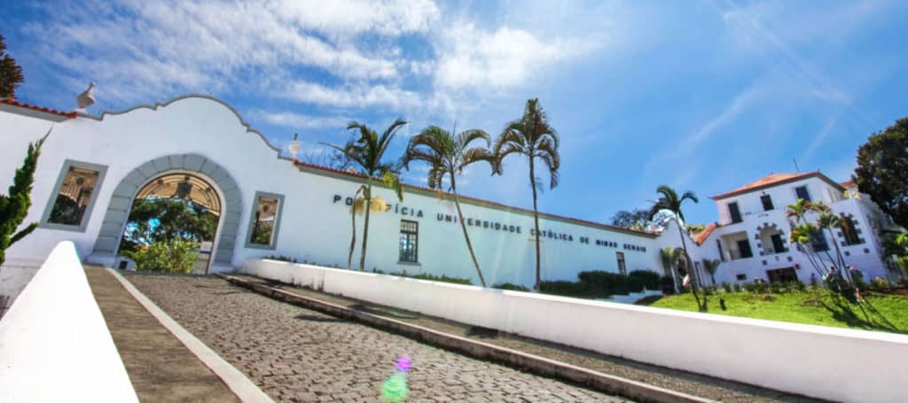 A imagem mostra a entrada principal e a fachada branca da Pontifícia Universidade Católica de Minas Gerais, em meio a palmeiras e um céu azul – um cenário clássico de BH pra universitários.