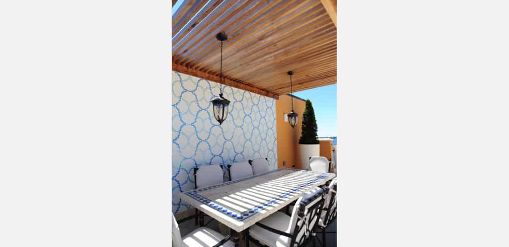 Área de jantar ao ar livre com mesa retangular, cadeiras acolchoadas, parede de destaque em papel de parede azul e branco, pérgola com teto de ripas de madeira e duas luminárias suspensas em estilo lanterna.