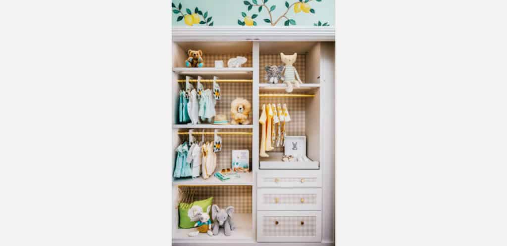 Um armário de bebê organizado com roupas penduradas, bichinhos de pelúcia, livros, cestos e gavetas. O fundo apresenta um charmoso papel de parede com tema de limão, e o armário tem uma decoração organizada e colorida.