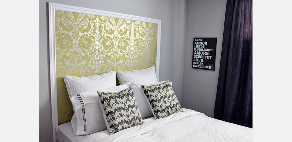 Uma cama impecavelmente arrumada, com roupa de cama branca e duas almofadas estampadas, repousa sobre uma cabeceira de papel de parede dourado e branco. Uma obra de arte emoldurada com a palavra "amor" em vários idiomas está pendurada na parede cinza ao lado.