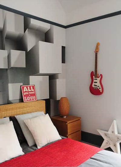 Um quarto moderno apresenta uma parede geométrica tridimensional atrás da cama, uma guitarra elétrica vermelha em exposição, uma placa com a frase "All you need is love" (Tudo o que você precisa é amor) e um criado-mudo com um abajur e decoração em forma de estrela.