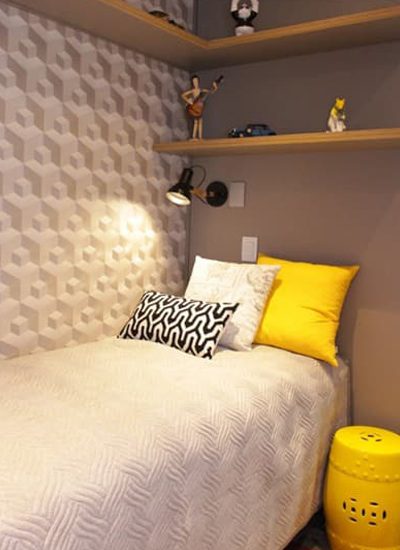 Um quarto aconchegante com uma cama de solteiro, roupa de cama branca texturizada, travesseiros estampados em preto e branco e amarelos, uma parede 3D geométrica em papel de parede cinza e branco, uma mesa lateral amarela e prateleiras de madeira com decoração acima da cama.