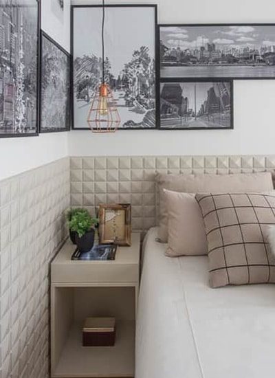 Um quarto moderno com uma cama bege, travesseiros xadrez e lisos, um pequeno criado-mudo bege com uma planta e livros, e fotos emolduradas em preto e branco na parede acima da cama, realçadas por um elegante papel de parede 3D para maior profundidade.