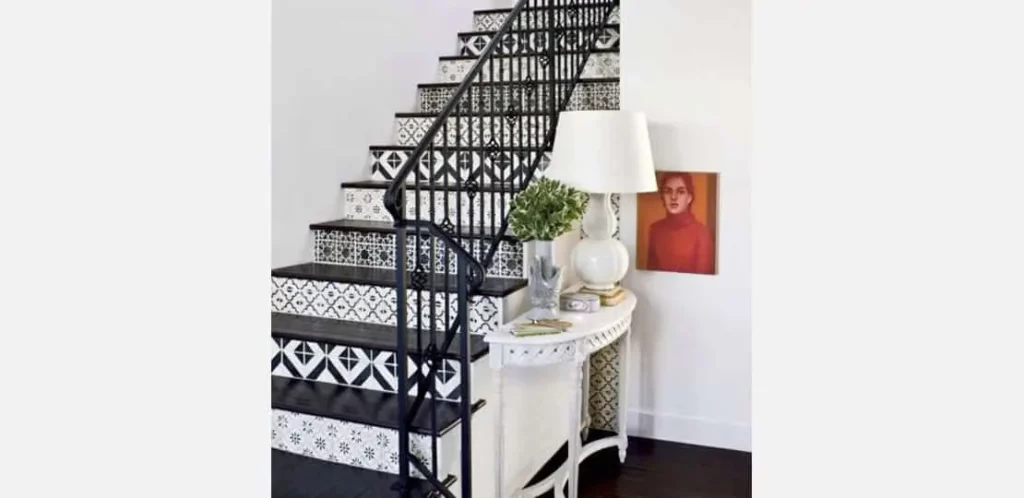 Uma escada com azulejos estampados em preto e branco, um corrimão de ferro preto e uma mesa de console branca abaixo dela, contendo um abajur, livros, uma planta e um retrato emoldurado encostado na parede com um elegante papel de parede como pano de fundo.