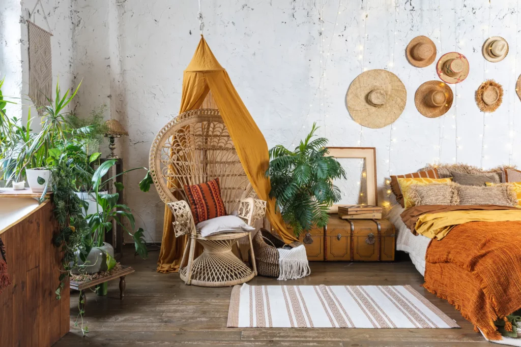 Foto de um quarto com decoração boho. O quarto tem chapéus de palha na parede, cadeira, plantas e uma cama confortável.
