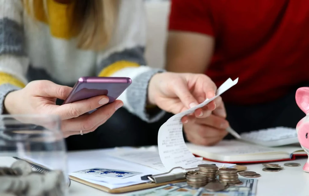 Imagem de duas pessoas sentadas em uma mesa contando moedas e dinheiro. Uma segura um telefone e a outra um recibo. Documentos, moedas e um cofrinho rosa estão sobre a mesa