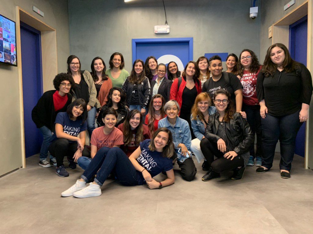 Um grupo de 21 pessoas, a maioria mulheres, posa e sorri em um corredor com portas azuis. Algumas estão em pé, outras ajoelhadas ou sentadas na frente. O clima amigável e descontraído reflete o espírito do Trabalhar no QuintoAndar.