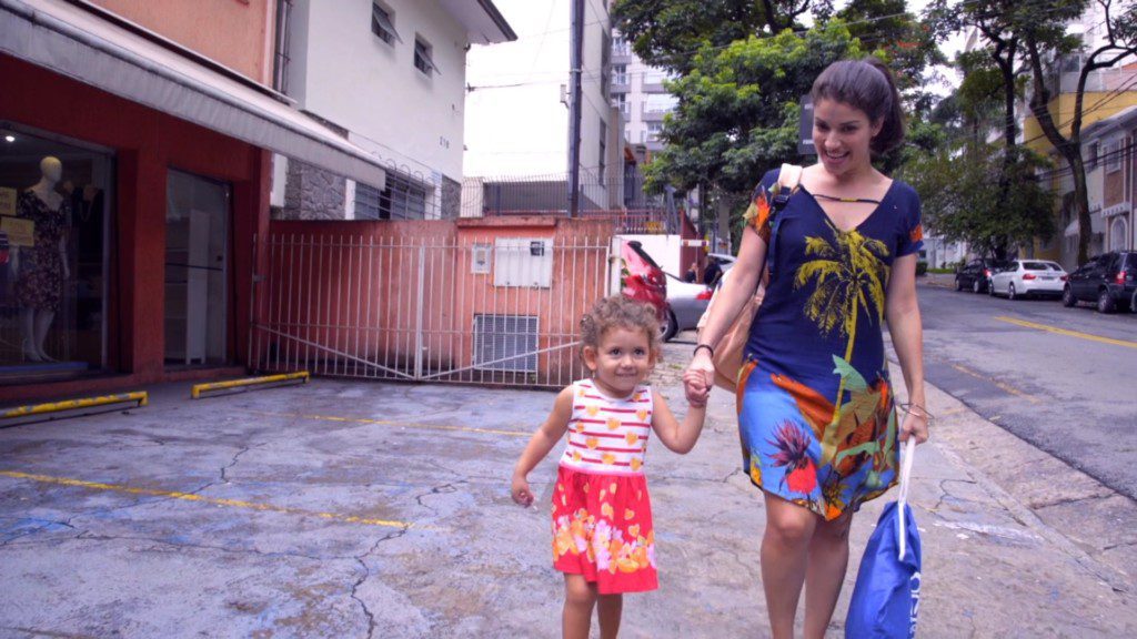 Uma mulher e uma jovem caminham de mãos dadas em um estacionamento urbano, abraçando as mudanças. A mulher carrega uma bolsa azul e ambas estão vestidas com roupas coloridas de verão. Prédios, carros estacionados e árvores são visíveis ao fundo.