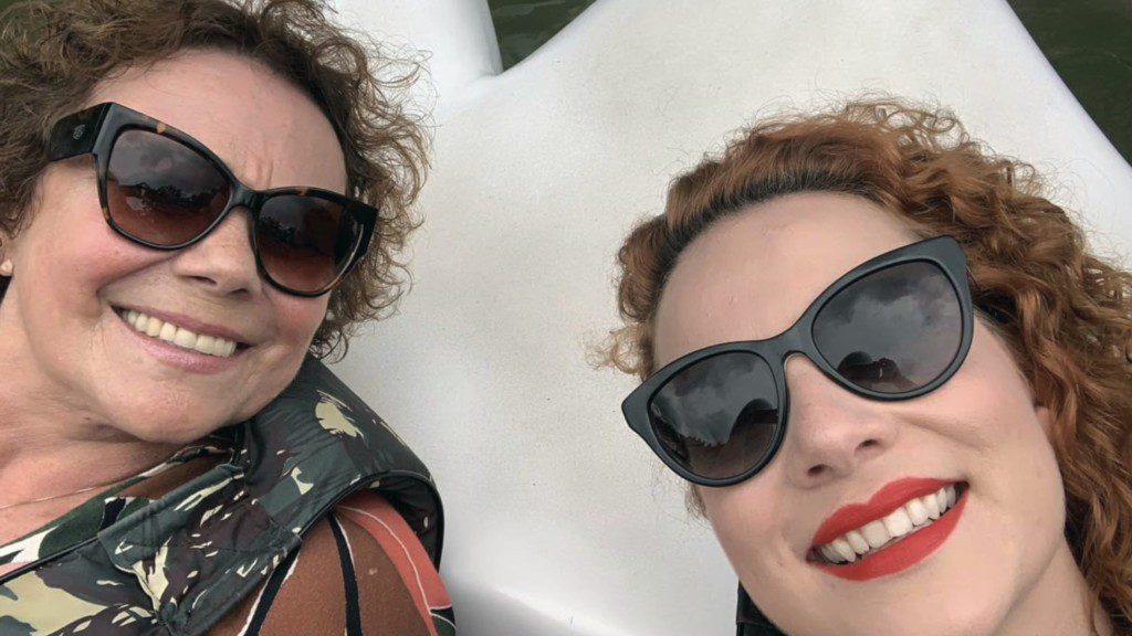 Duas mulheres de cabelos cacheados e óculos escuros sorriem para uma selfie ao ar livre, apreciando a arte do morar bem. Uma usa uma jaqueta estampada, a outra, um batom vermelho vibrante — elas relaxam juntas em uma superfície branca, irradiando estilo e felicidade.