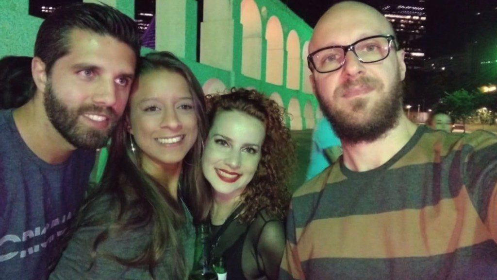 Quatro adultos posam juntos para uma selfie à noite em frente a um prédio iluminado com arcos. Sorrindo e segurando uma bebida, eles personificam a alegria de Morar bem em meio às luzes da cidade ao fundo.