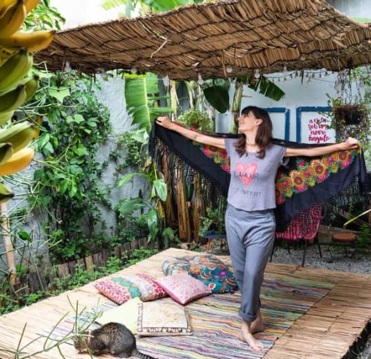 Uma mulher está descalça sobre um tapete colorido em um pátio externo exuberante, esticando bem o xale. O cenário vibrante, com almofadas e tapetes decorativos, evoca inspiração de decoração de quarto de crianças como um gato descansando sob uma cobertura rústica de bambu.