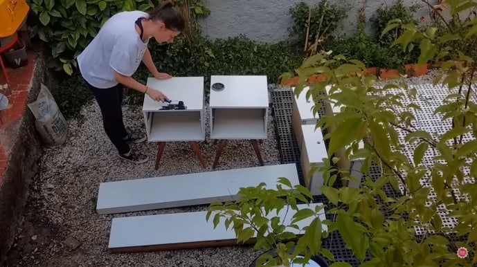 Uma mulher está ao ar livre, pintando ou montando dois pequenos armários brancos apoiados sobre pernas — perfeitos para decoração de quarto infantil. Várias tábuas de madeira branca estão próximas, cercadas por plantas verdejantes.