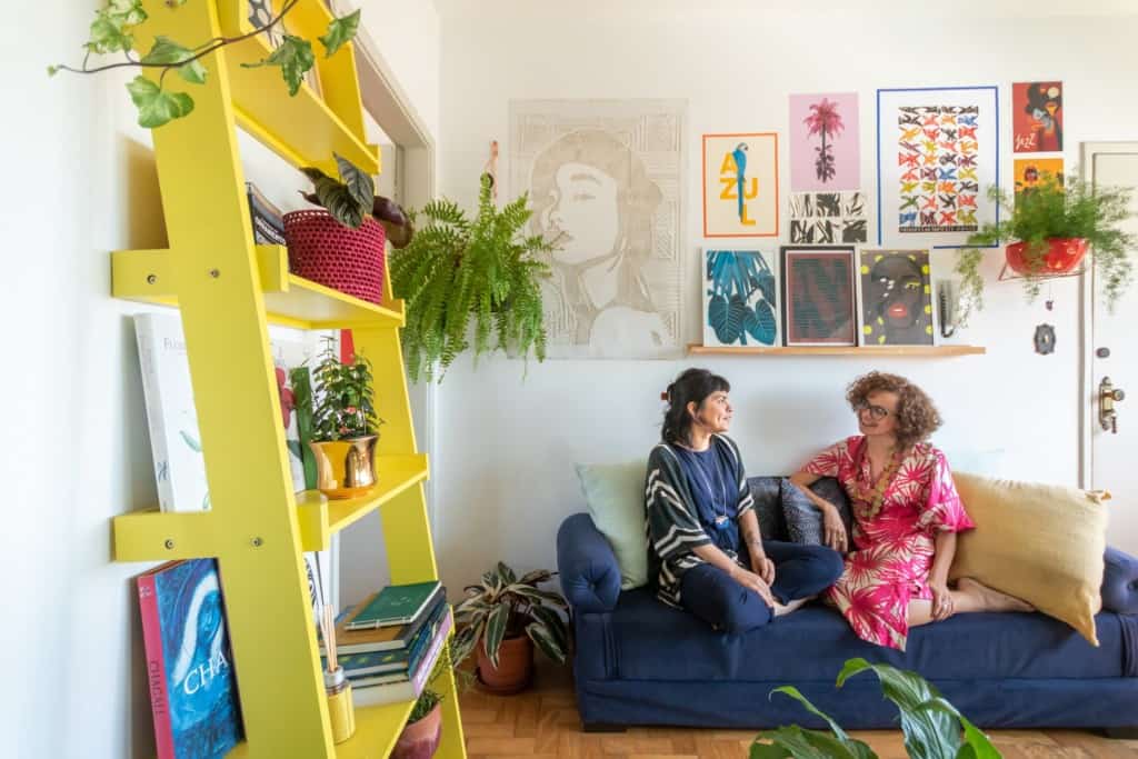 Duas mulheres conversam sentadas em um sofá azul em uma sala de estar iluminada e repleta de plantas. Arte e plantas coloridas decoram as paredes, enquanto uma estante amarela com livros e plantas contribui para a sensação de liberdade e criatividade no espaço.