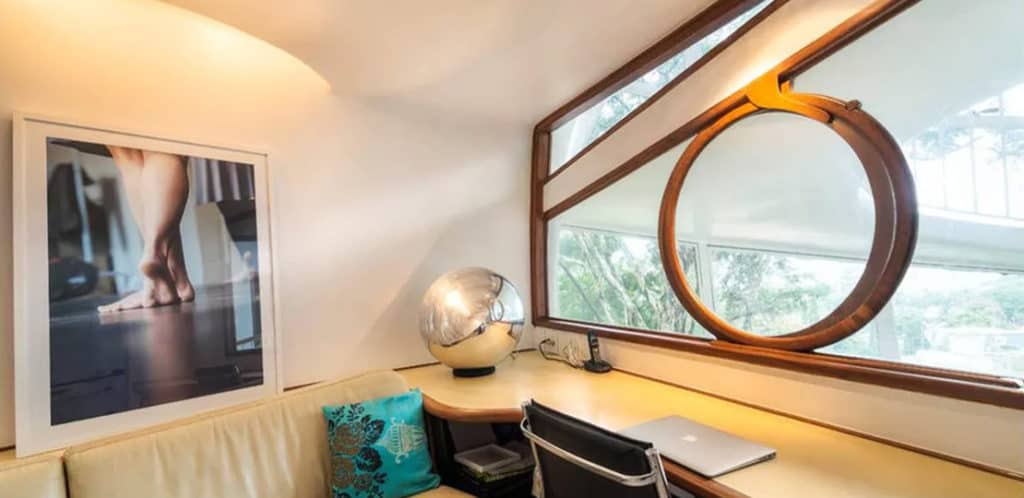 Um home office moderno com mesa de madeira, laptop, cadeira preta, janela redonda e luminária globo prateada oferece o ambiente perfeito para qualquer Stalker de imóvel. O sofá bege com almofada azul fica abaixo de uma grande foto de pés na parede.