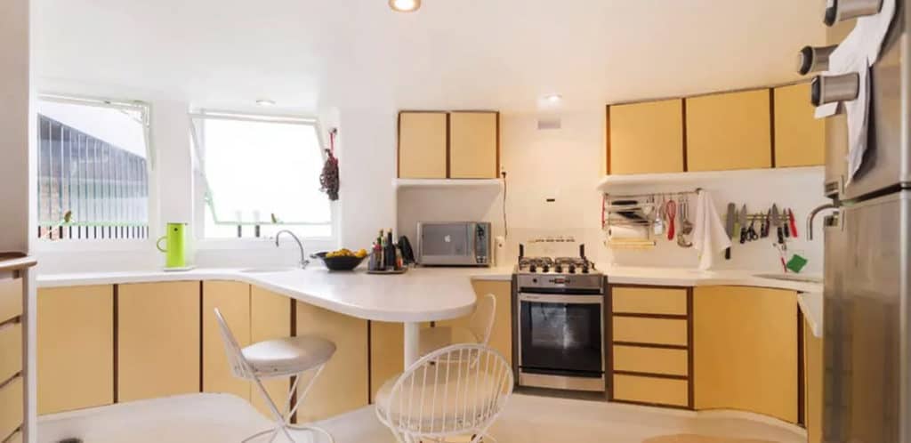 Uma cozinha moderna com armários amarelo-claros, bancada branca curva, fogão a gás, eletrodomésticos de aço inoxidável, duas cadeiras e janelas amplas que permitem a entrada de luz natural — perfeita para qualquer Stalker de imóvel em busca de funcionalidade e estilo. Utensílios de cozinha pendurados acima do balcão.