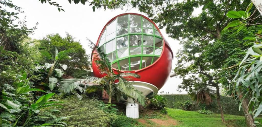 Uma casa moderna, vermelha e branca, em formato de cápsula, com grandes janelas curvas, fica no alto, entre árvores verdejantes — um sonho tornado realidade para qualquer Stalker de imóvel que esteja explorando o refúgio perfeito na encosta.
