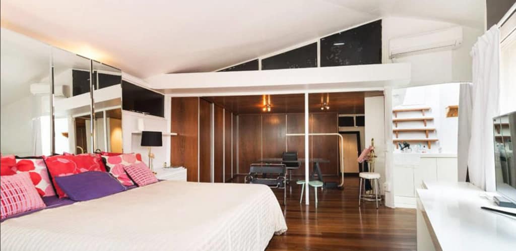 Um quarto espaçoso com piso de madeira, uma cama grande com almofadas coloridas, armários espelhados, uma TV na parede e um espaço de trabalho moderno — ideal para qualquer Stalker de Imóveis em busca de estilo. A luz natural entra por uma janela com cortinas brancas.