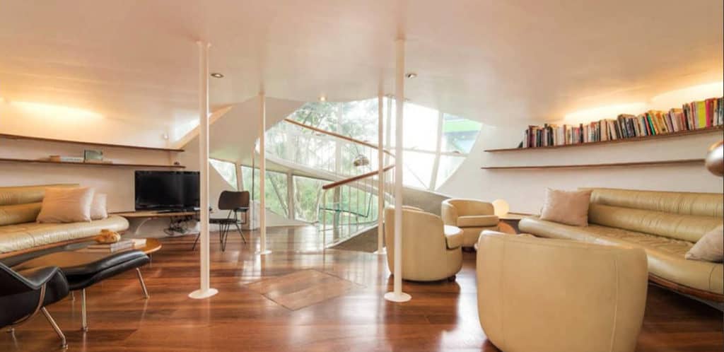 Sala de estar moderna com sofás de couro bege, poltronas, piso de madeira, TV, estantes com livros — exatamente o tipo de espaço que um Stalker de imóvel adoraria — além de grandes janelas inclinadas que oferecem luz natural e vista para as árvores.