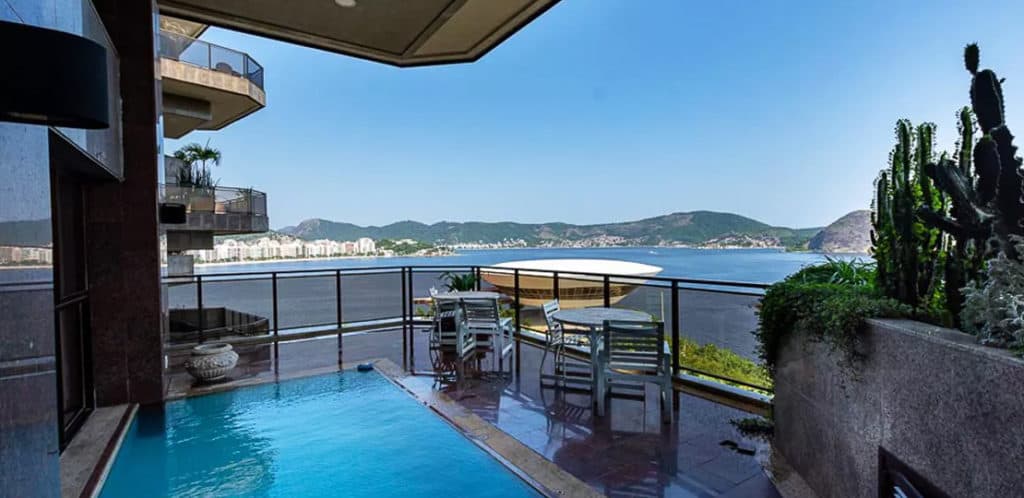 Um pátio na cobertura com uma pequena piscina, mesas e cadeiras ao ar livre, plantas em vasos e um corrimão de vidro tem vista para uma baía pitoresca e colinas distantes — um verdadeiro sonho de Stalker de imóvel sob um céu azul claro.