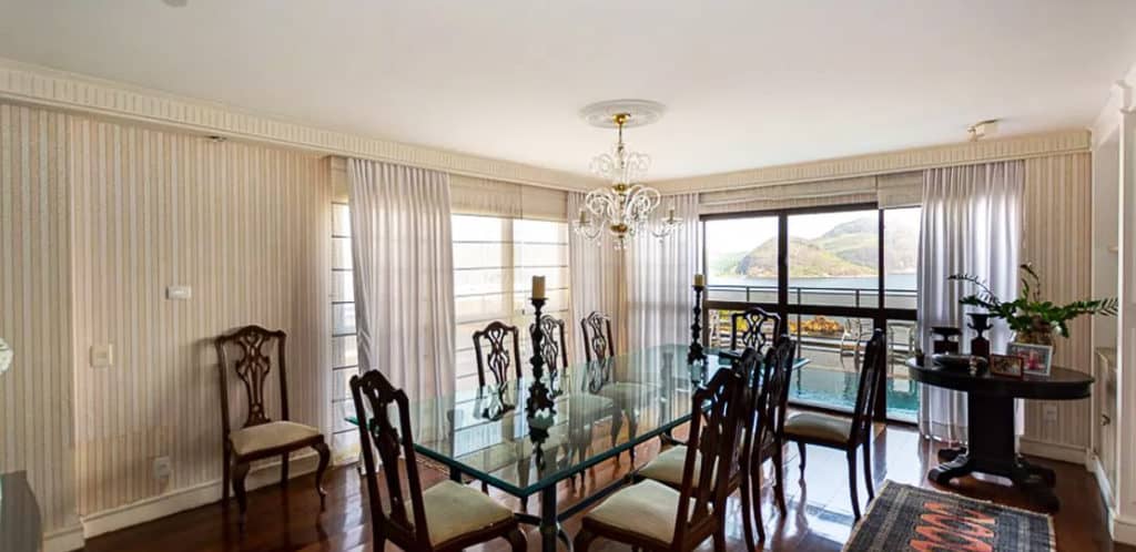 Sala de jantar elegante com mesa de vidro e oito cadeiras de madeira, lustre acima, janelas amplas com cortinas transparentes que deixam entrar luz natural — perfeita para qualquer aventureiro em busca de vista para as montanhas. Itens decorativos ficam em mesas laterais elegantes.