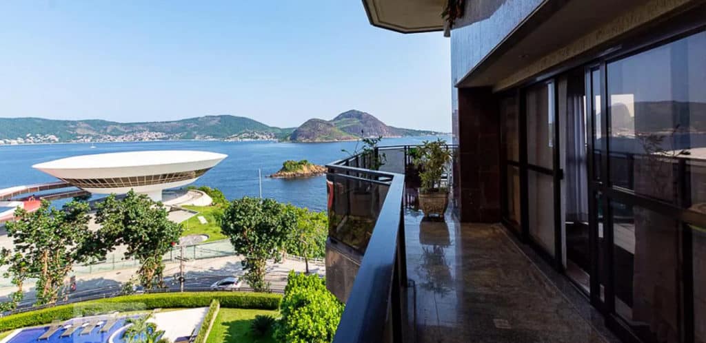 Uma varanda espaçosa com grades de vidro tem vista para uma baía, colinas verdejantes e o moderno Museu de Arte Contemporânea de Niterói, em formato de disco, no Rio de Janeiro — um achado ideal para qualquer perseguidor de imóvel em um dia claro e ensolarado.
