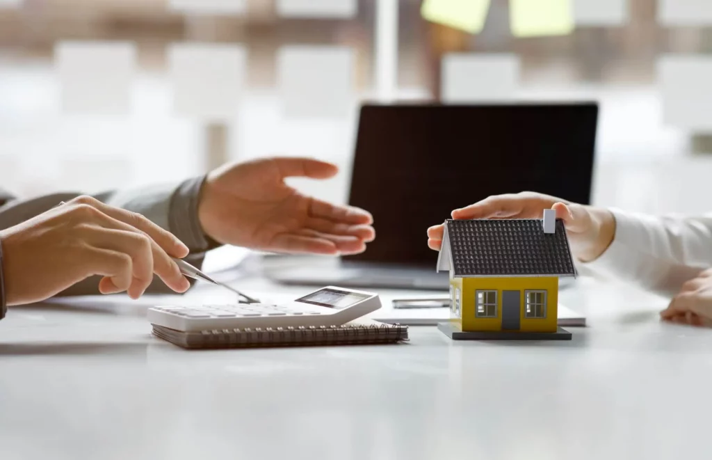 Imagem de uma pessoa apontando para uma calculadora enquanto outra pessoa segura uma casa de madeira em tamanho miniatura em cima de uma mesa para ilustrar matéria sobre se vale a pena financiar imóvel
