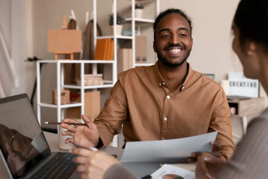 Imagem de um homem sorridente, vestindo uma camisa marrom, aponta para a tela de um notebook, enquanto discute documentos e o processo de como vender uma casa com uma mulher