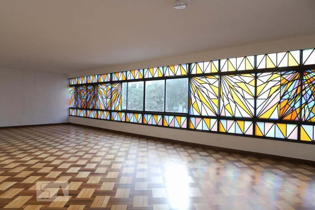 Sala espaçosa e vazia, com piso de madeira e grandes janelas com vitrais geométricos coloridos, permitindo a entrada de luz natural — perfeita para ouvir Duda Beat enquanto a luz do sol dança no chão.