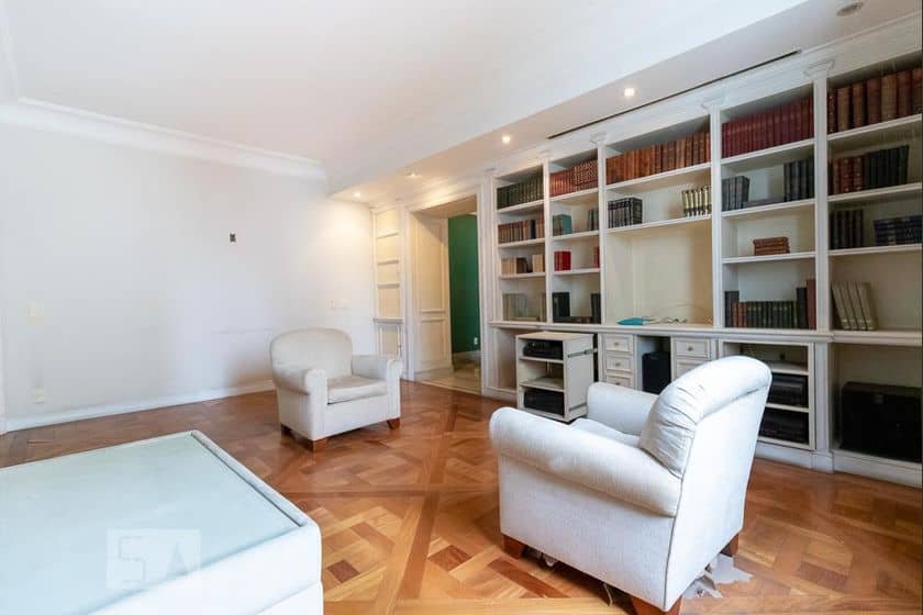 Uma sala espaçosa e iluminada com piso de parquet, duas poltronas estofadas, uma mesa de centro de vidro e uma grande estante embutida cheia de livros e decorações inspiradas em Duda Beat ao longo de uma parede.