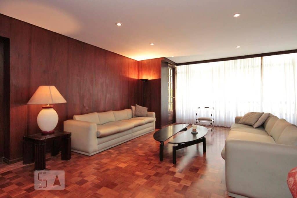 Uma sala de estar espaçosa com paredes revestidas de madeira, piso de parquet, dois sofás creme e uma mesa de centro de vidro cria um ambiente aconchegante, perfeito para relaxar ao som das músicas de Duda Beat, enquanto a luz natural entra pelas grandes janelas com cortinas brancas transparentes.