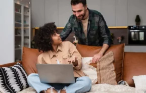 Imagem de uma mulher sentada em um sofá com um laptop no colo, discutindo os serviços digitais da Imobiliária com um homem atrás dela. Ambos parecem estar conversando em sua sala de estar moderna e aconchegante.