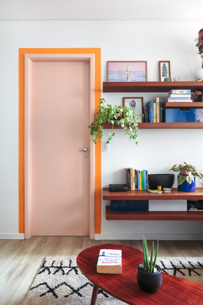 Um moderno apartamento de 1 dormitório em Pompeia, com uma porta rosa-claro emoldurada por prateleiras de madeira flutuantes em laranja com livros e plantas, uma pequena mesa de centro vermelha e um tapete com estampa clara sobre piso de madeira.