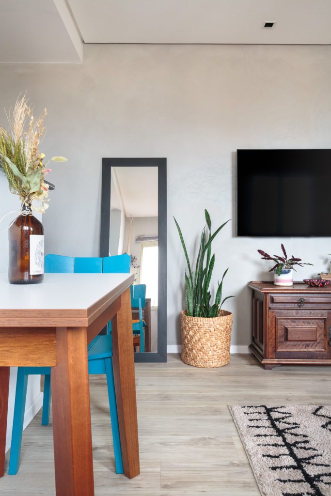 Uma sala de estar moderna em um apartamento de 1 dormitório em Pompeia apresenta piso de madeira clara, uma mesa de madeira com cadeiras azuis, um grande espelho de chão, planta em vaso, TV de parede, armário de madeira, tapete estampado e flores secas em uma garrafa de vidro marrom.