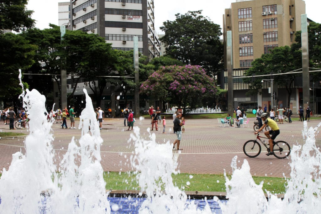 Pessoas se reúnem em uma praça com árvores e prédios altos — como os próximos ao Apartamento Lourdes BH — ao fundo. Em primeiro plano, a água de uma fonte jorra para cima enquanto várias pessoas caminham e alguém anda de bicicleta perto do centro.
