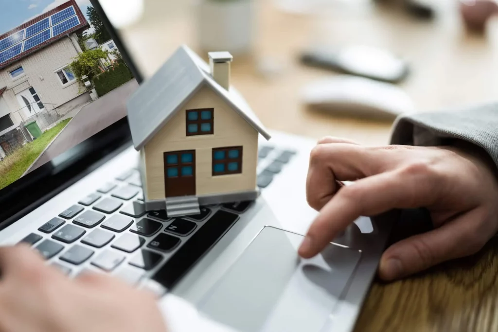 Imagem de uma pessoa olhando a foto de um imóvel no notebook enquanto uma casa de madeira em tamanho miniatura está em cima do teclado para ilustrar matéria sobre como vender mais imóveis pela internet