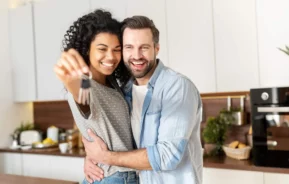 Imagem de um casal formado por um homem e uma mulher se abraçando na cozinha de casa enquanto a mulher segura a chave de um imóvel para ilustrar matéria sobre como vender rápido um imóvel