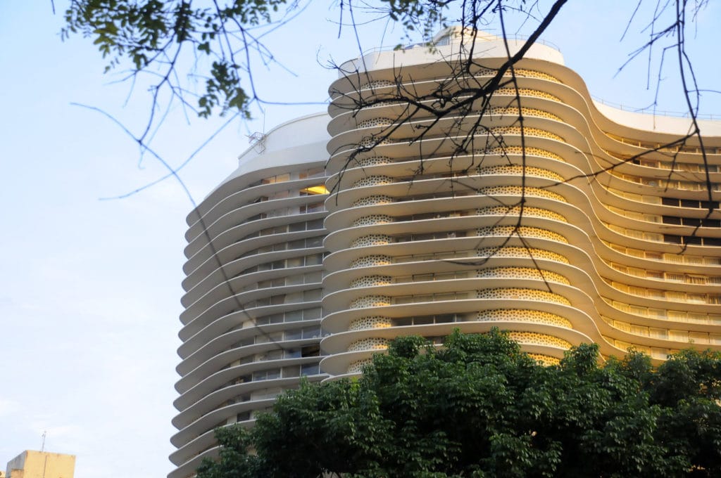 Um arranha-céu moderno e curvo com amplas varandas e uma fachada bege é iluminado pela luz do sol, parcialmente obscurecida por árvores frondosas — uma visão elegante no Apartamento Lourdes BH.