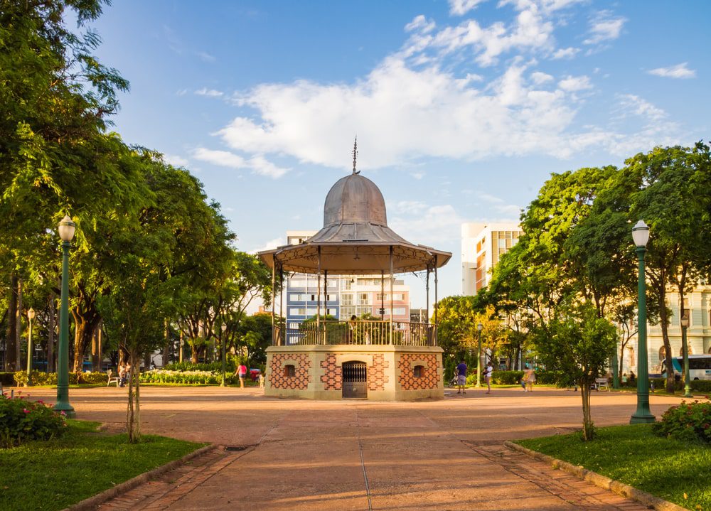 Um gazebo de metal fica no centro de um parque cercado por árvores, postes de luz e caminhos, com pessoas caminhando e edifícios modernos — como os próximos ao Apartamento Lourdes BH — visíveis ao fundo sob um céu parcialmente nublado.